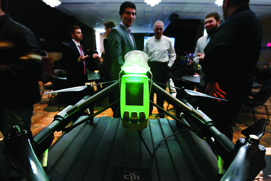 A green camera is mounted on a black drone with people in the background.