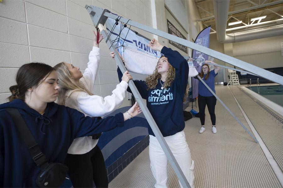 A group of students work together to put up a large sign on a pool deck.